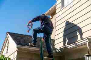 Middle aged handyman getting off of a green ladder to hang a string of red and white colored Christmas lights.